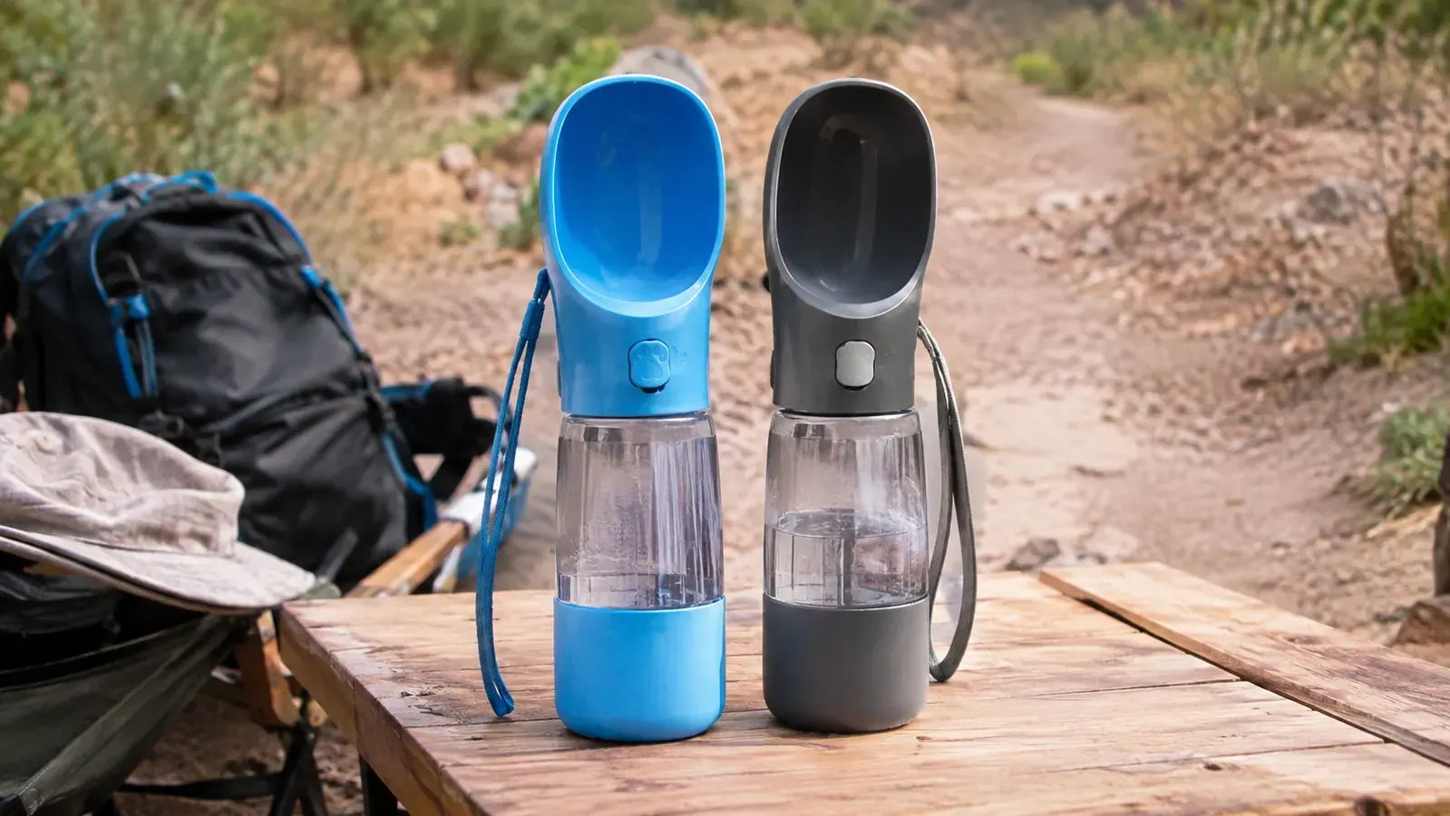 Two portable dog water bottles side by side on a wooden camp table, one blue and one charcoal, with a trail in the background