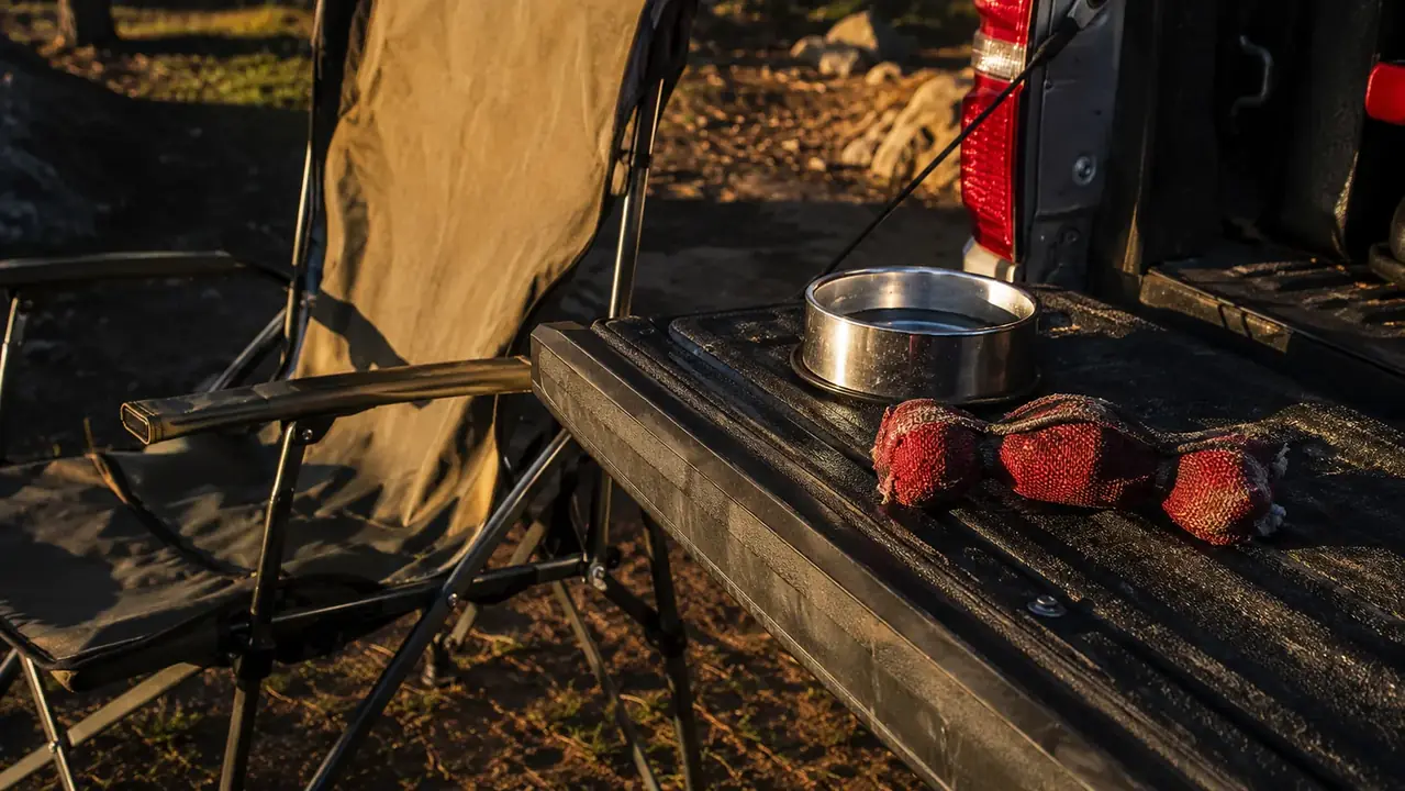 A chewed-in Nylabone Power Chew toy resting on a truck tailgate beside a camp chair and a metal water dish at golden hour